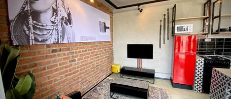 Un soggiorno moderno con parete in mattoni, televisione a schermo piatto, frigorifero rosso e divano nero con cuscini decorati.
