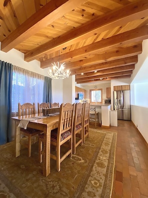 A dining area with a wooden table and chairs, a chandelier, and a kitchen in the background.