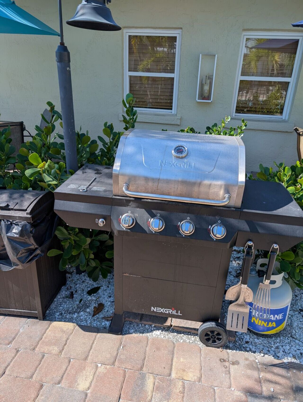 A NEXGRILL barbecue grill with four burners, a built-in thermometer, and a side burner. There are grilling tools and a trash can nearby.