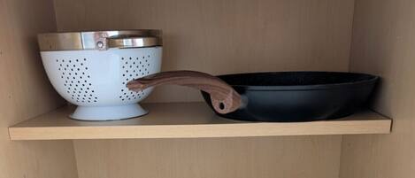 A kitchen shelf with various cookware including a colander, a frying pan, and several pots.