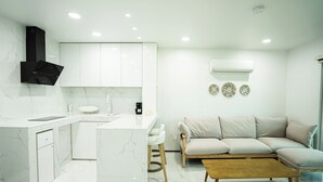 A modern kitchen with white cabinetry, a marble countertop, and a built-in stove. There is a beige sofa and a wooden coffee table in the living area. - Mariana Suites (Saipan)