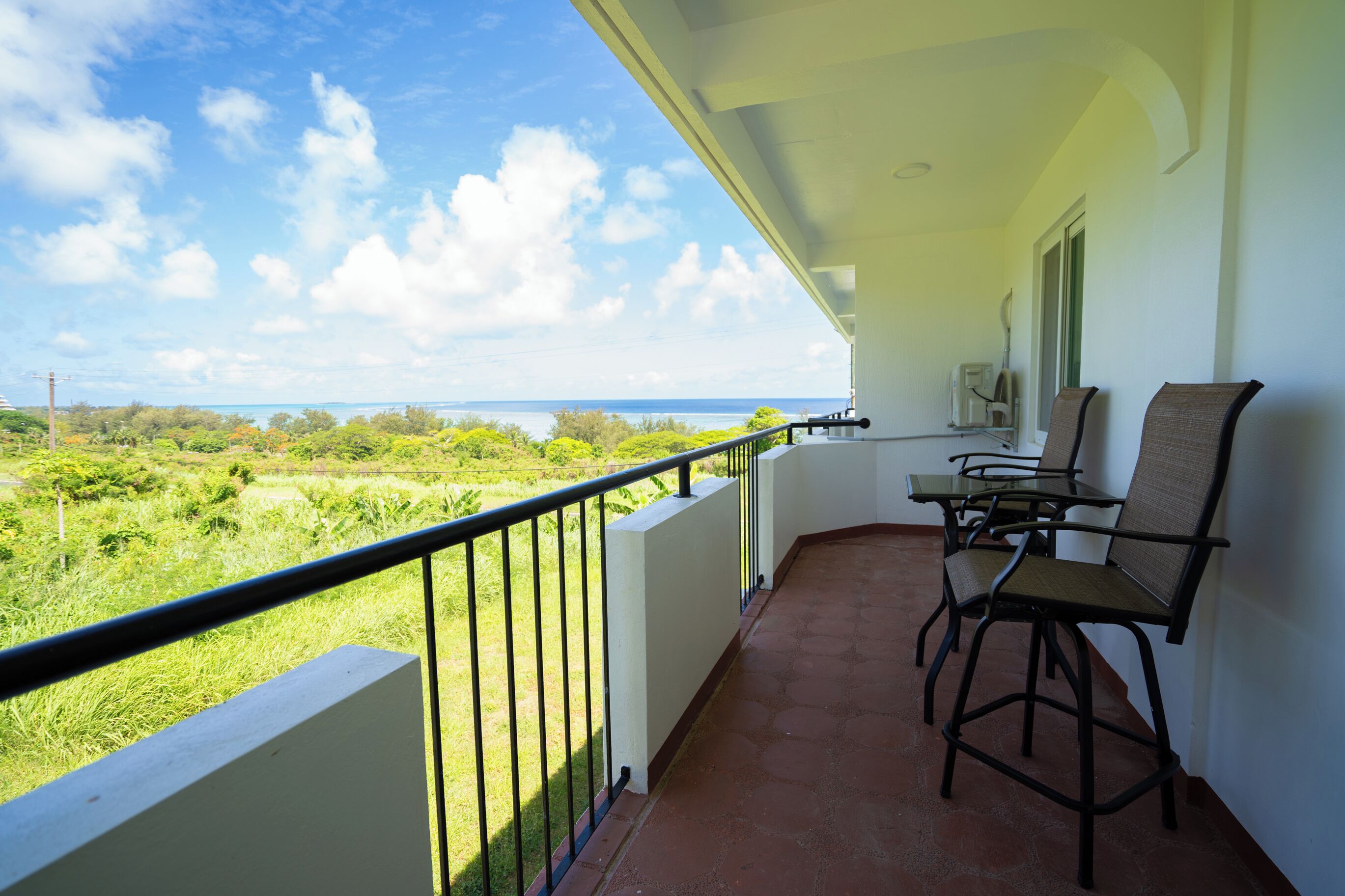 A balcony with a view of a grassy landscape and the sea. There are two chairs and a table on the balcony.