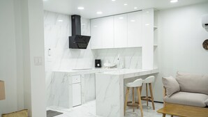 A modern kitchen with white marble countertops, a built-in stove, and a sink. There is a wooden table and a beige sofa in the living area. - Mariana Suites (Saipan)