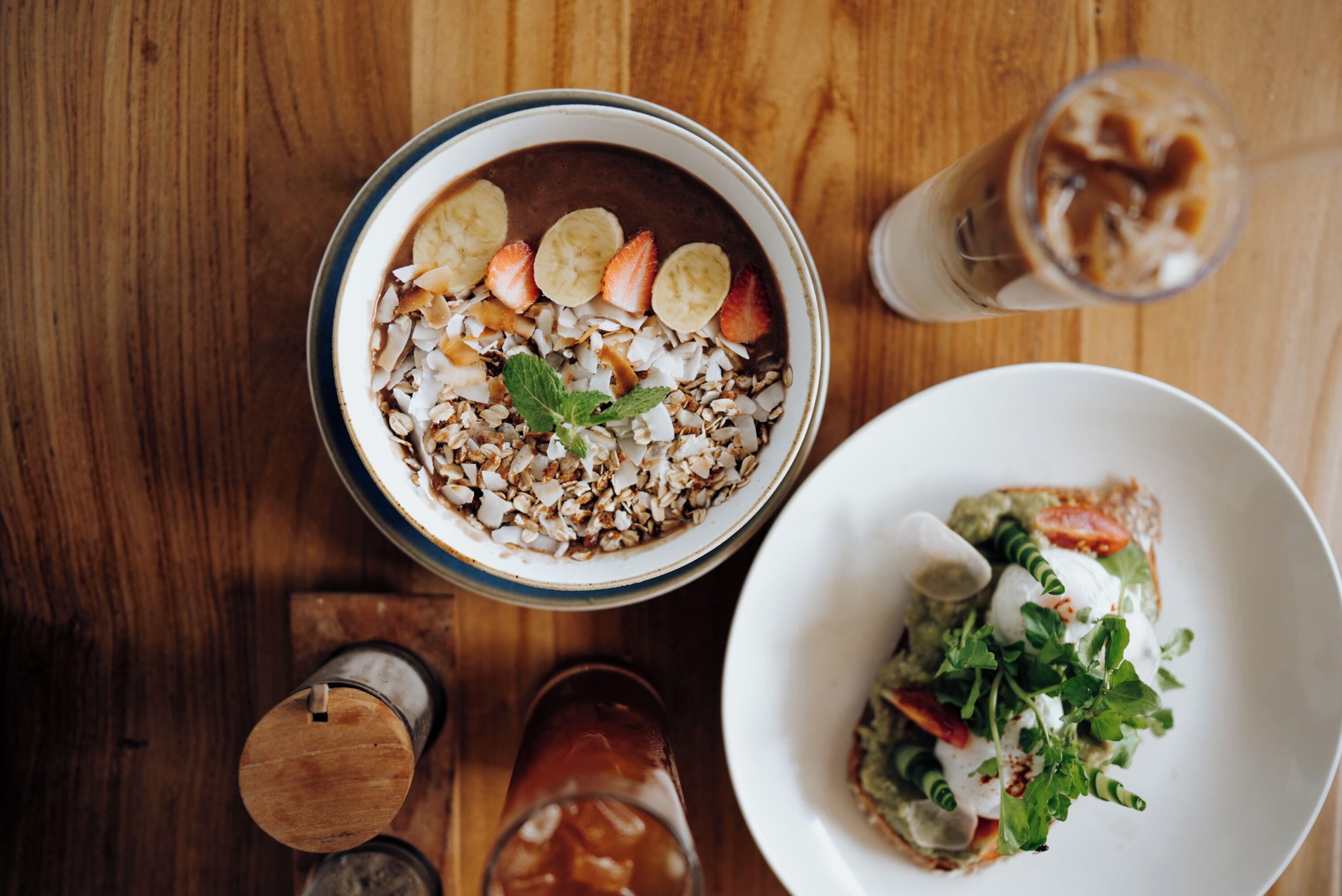 A dining table with a bowl of oatmeal topped with fruits and nuts, a glass of iced coffee, and a plate of salad.