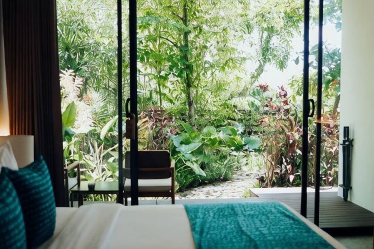 Una habitación con cama, una silla y vistas a un jardín exuberante a través de amplios ventanales.