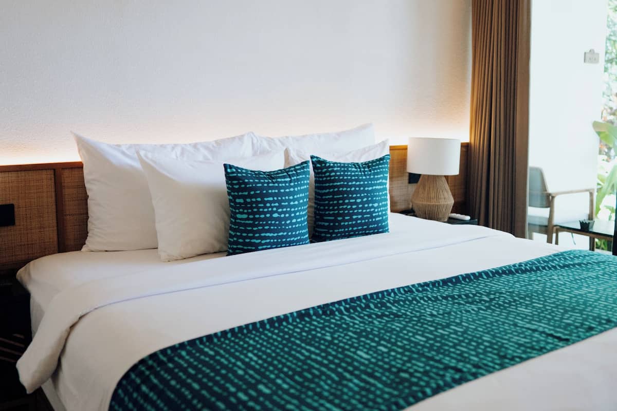 A hotel room with a large bed, a nightstand, a lamp, and a view of greenery outside.