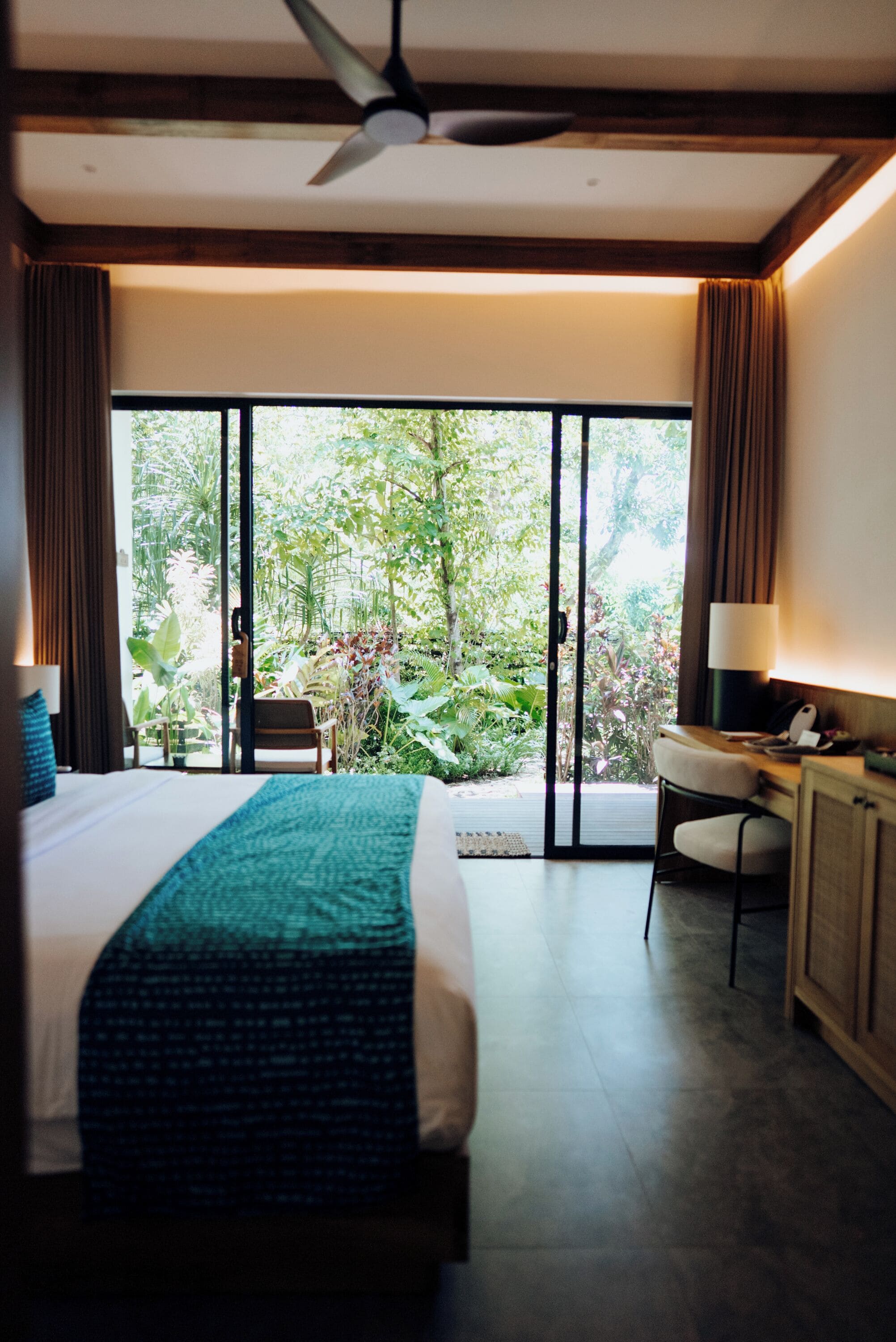 A hotel room with a bed, a desk, a chair, and a view of greenery through large windows.