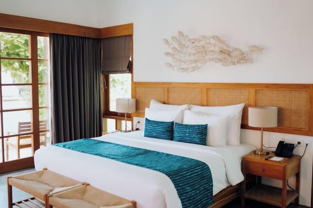 A hotel room with a large bed, a wooden headboard, a bench, a nightstand with a lamp, and a wall-mounted artwork.