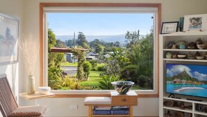 Ein Zimmer mit großem Fenster mit Blick auf eine grüne Landschaft, ein Bücherregal mit diversen Gegenständen und einem Fernseher, der eine tropische Szene zeigt.