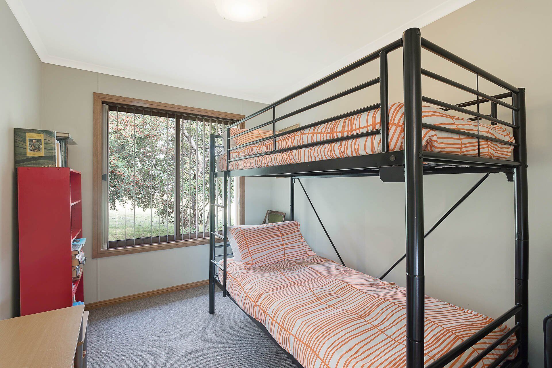 A bunk bed with striped bedding in a room with a window, a red bookshelf, and a small table.