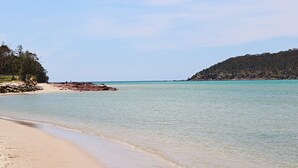 Ein Strand mit klarem türkisblauem Wasser, Sandküste und einer bewaldeten Hügelkette im Hintergrund.