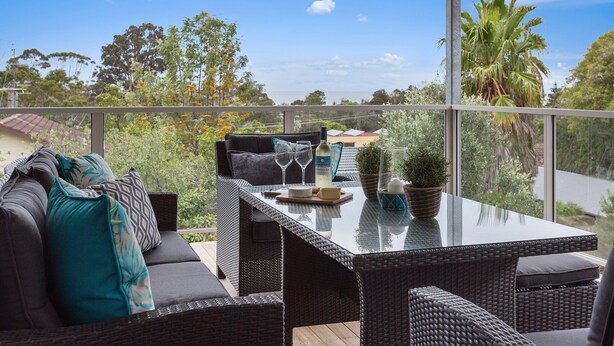 Ein Balkon mit Korbmöbeln, einem Glastisch und Blick auf Bäume und Gebäude.