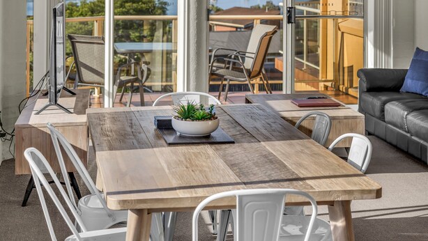 A modern dining area with a wooden table, white chairs, and a view of a lake with boats. - Baywatch Apartments Merimbula- 2 bedroom with beautiful views (Merimbula)