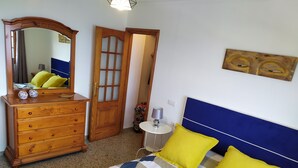 Een slaapkamer met een bed, een dressoir, een spiegel, een deur en een tafeltje met een lamp.