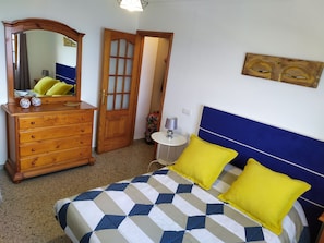 A bedroom with a bed, a dresser, a mirror, a door, and a small table with a lamp.