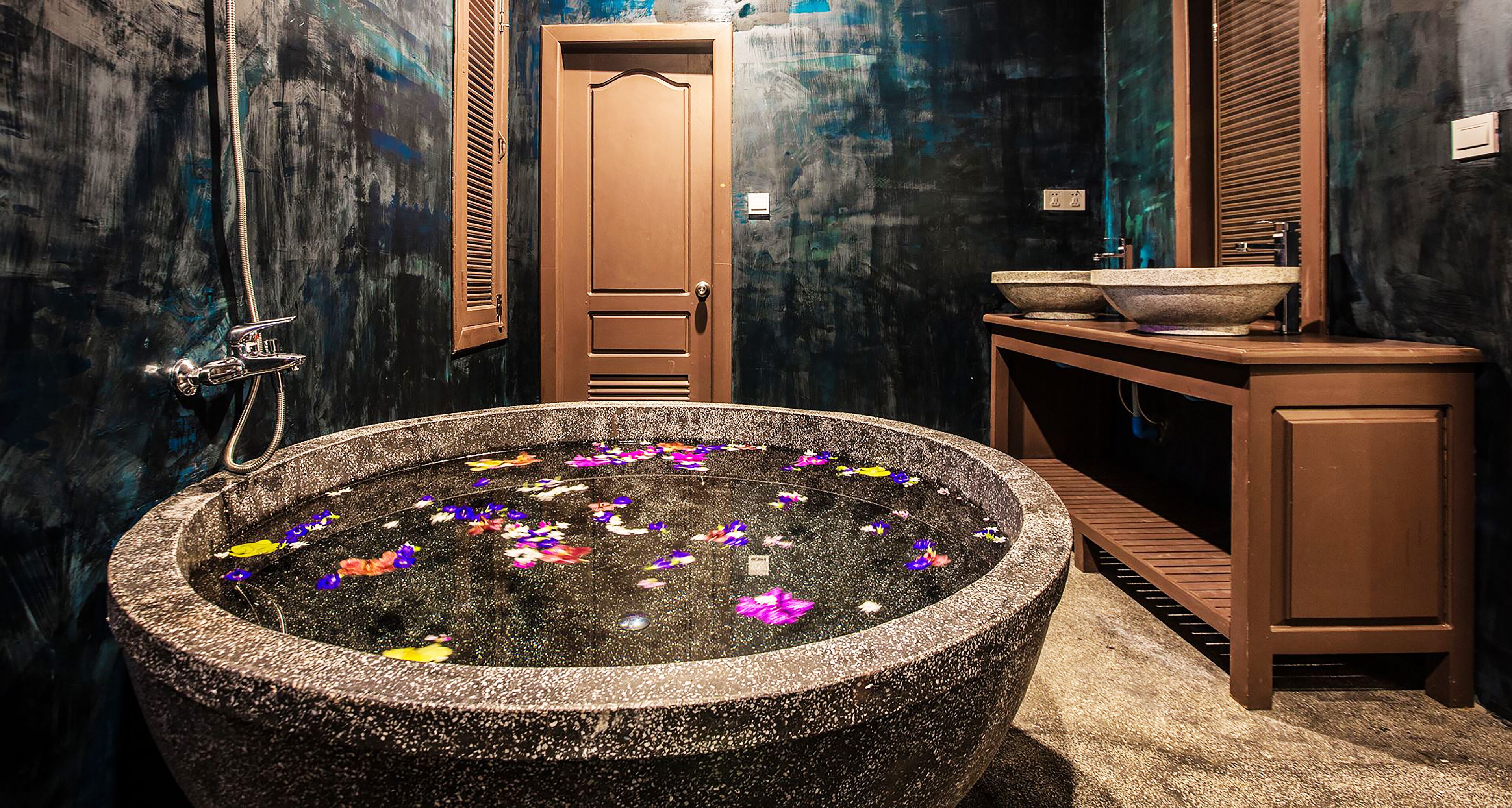 A bathroom with a freestanding bathtub filled with water and floating flowers, a wooden door, and a sink with a stone basin.
