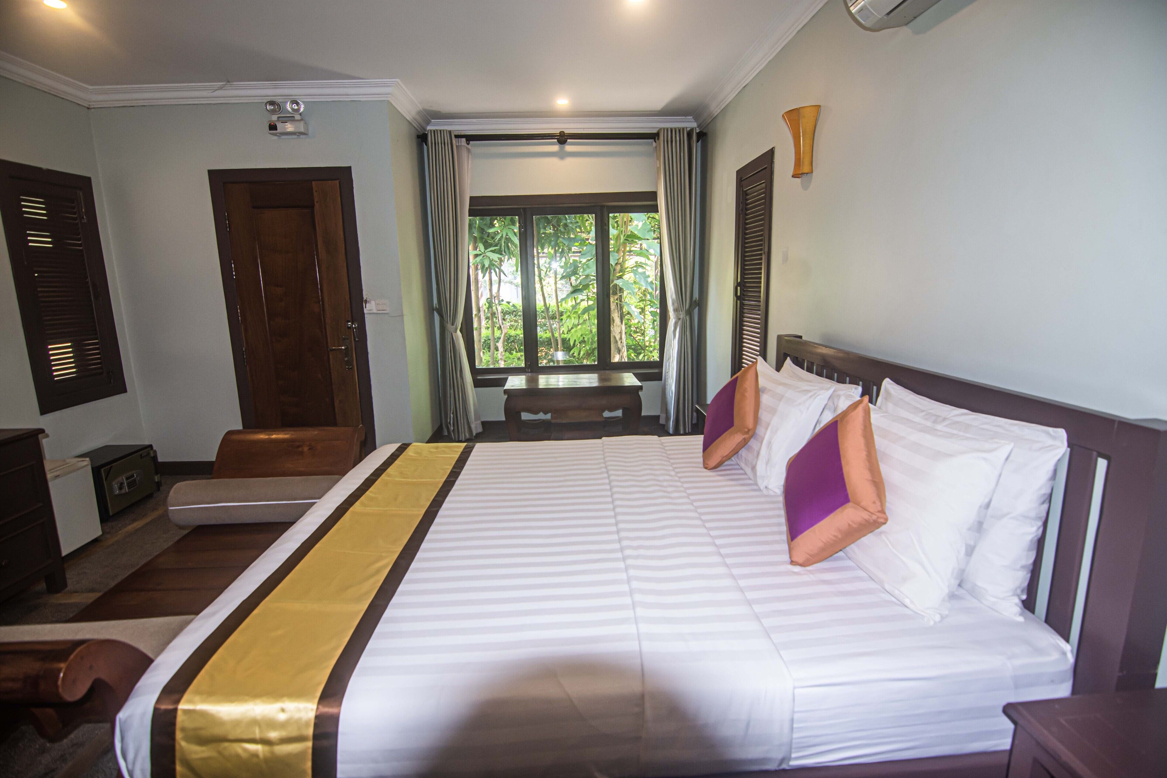 A hotel room with a large bed, a wooden bench, a desk, and a window with curtains.