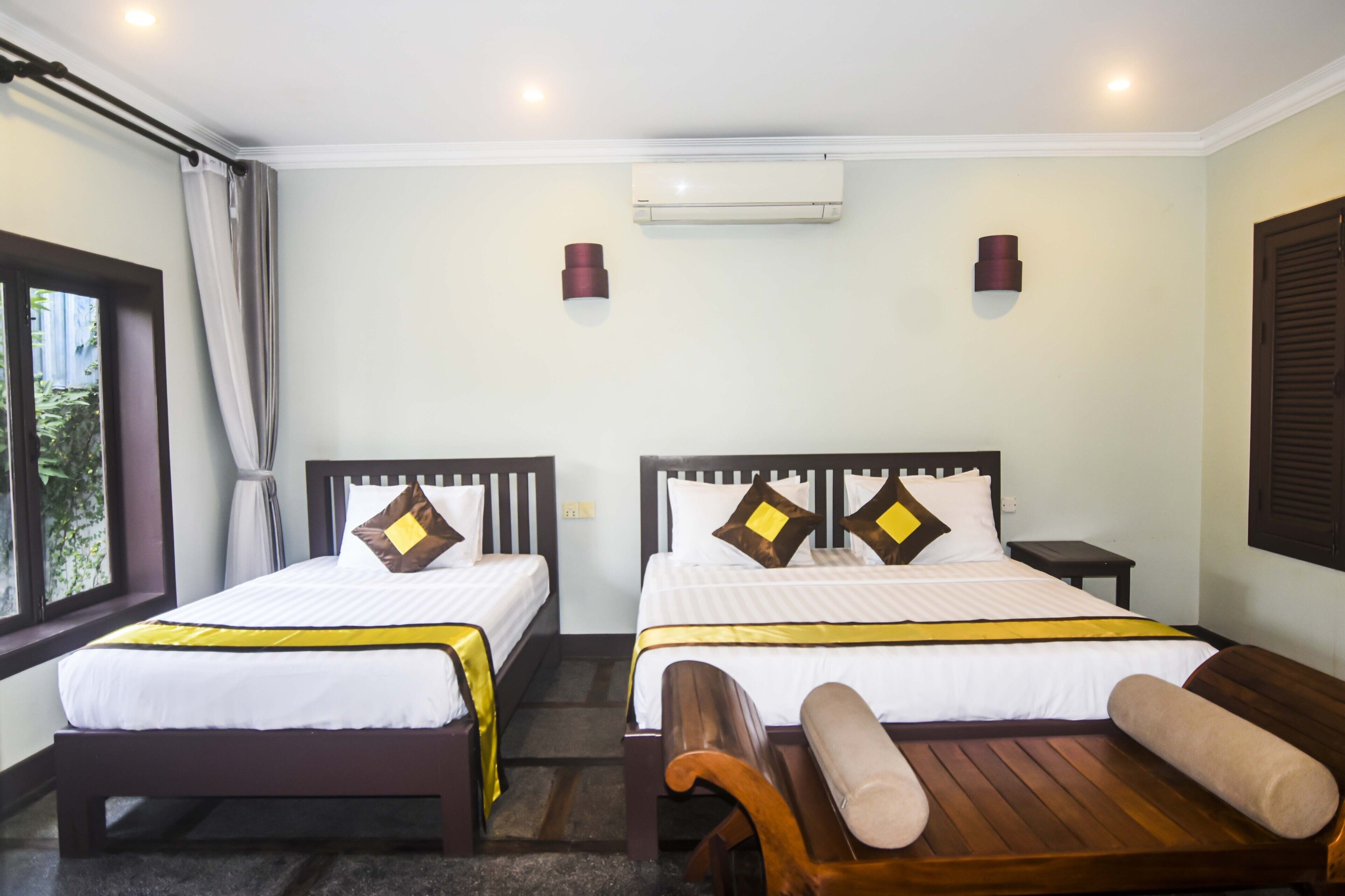 A hotel room with two beds, a wooden bench, an air conditioning unit, and a window with curtains.