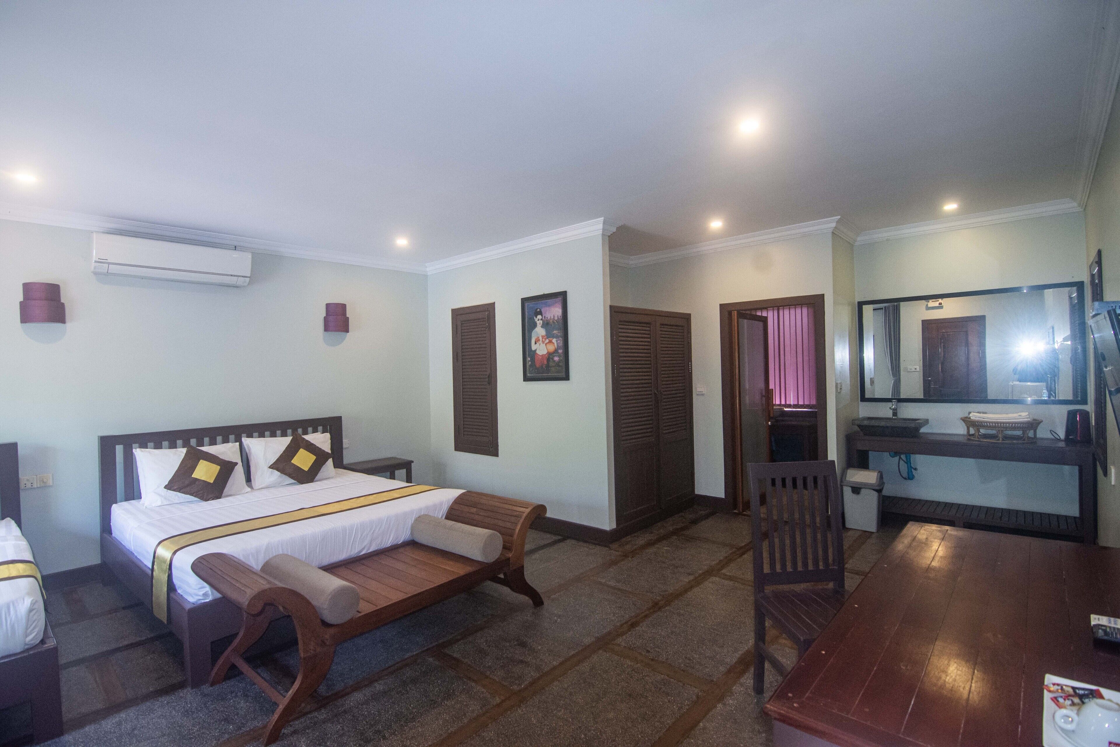 A hotel room with a bed, bedside table, bench, wooden floor, and a small table with a chair.