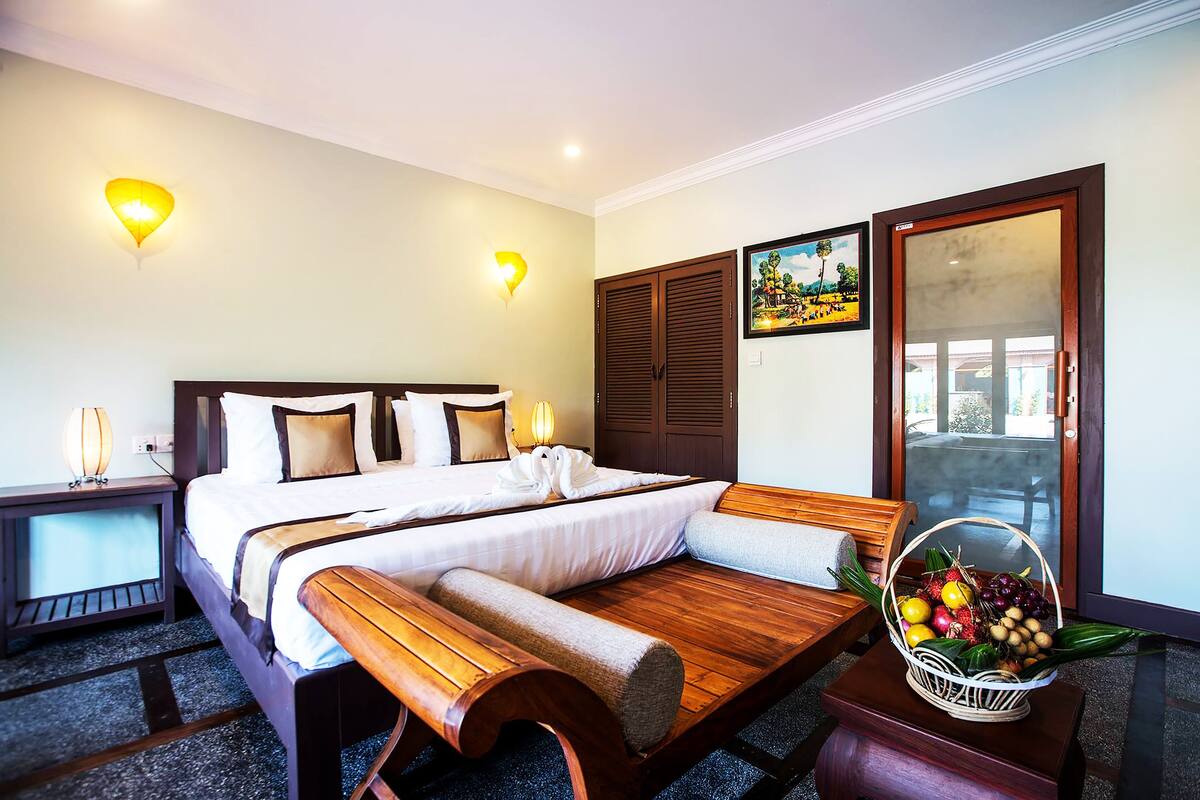 Quarto de hotel com cama, mesas de cabeceira, um banco de madeira, uma cesta de frutas e uma porta com vista.
