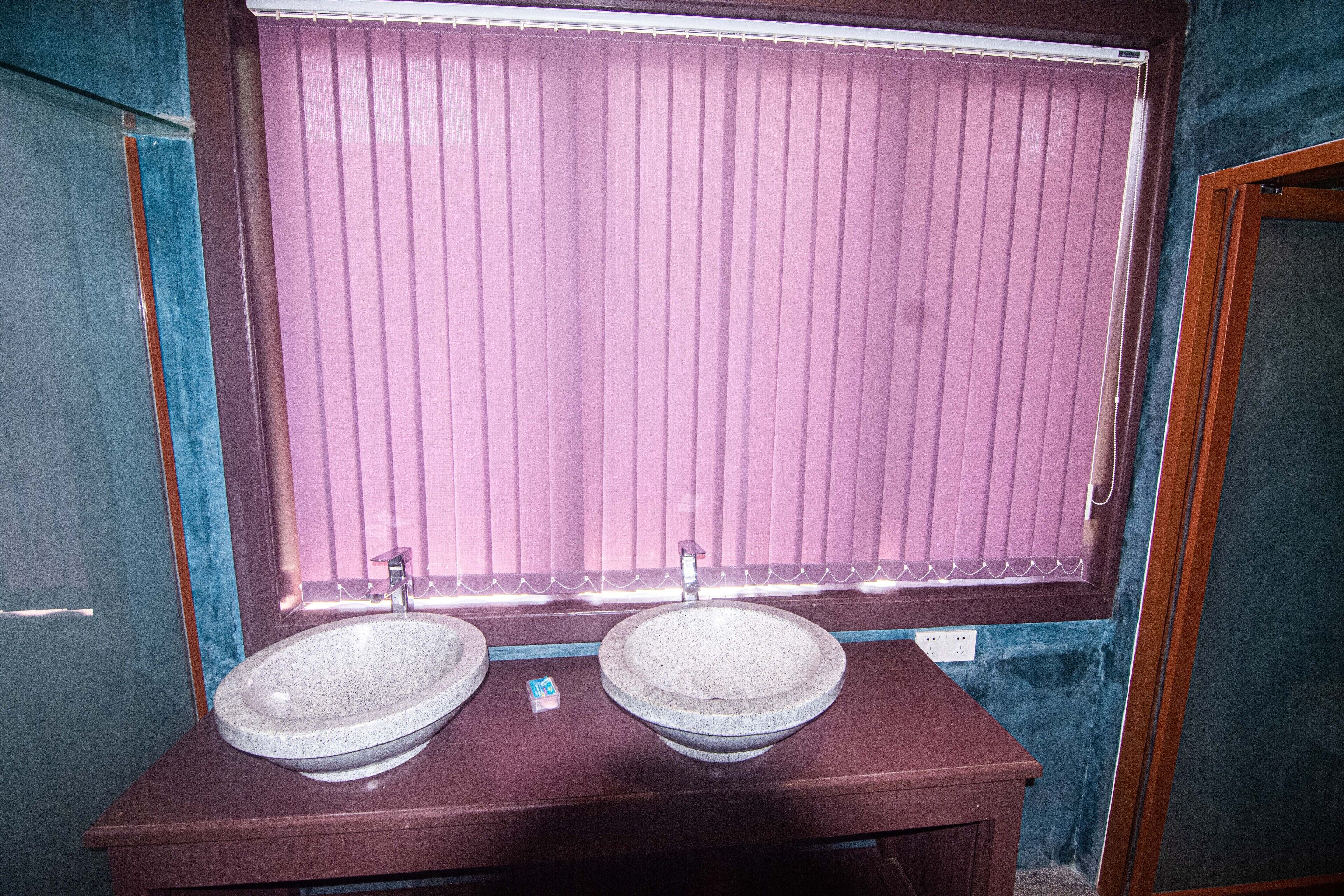 A bathroom with two stone sinks, a wooden vanity, and a window with pink blinds.