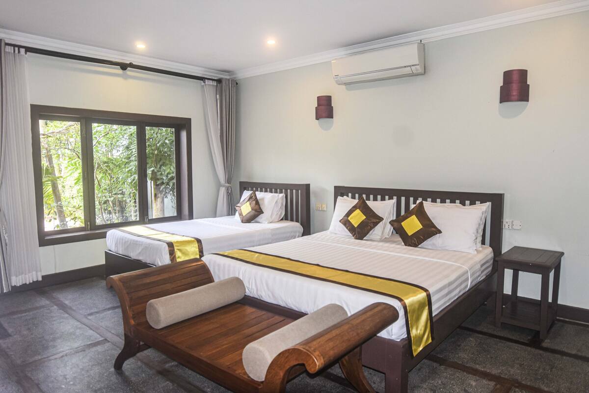 Quarto de hotel com duas camas, um banco, uma mesinha e uma janela com cortinas.