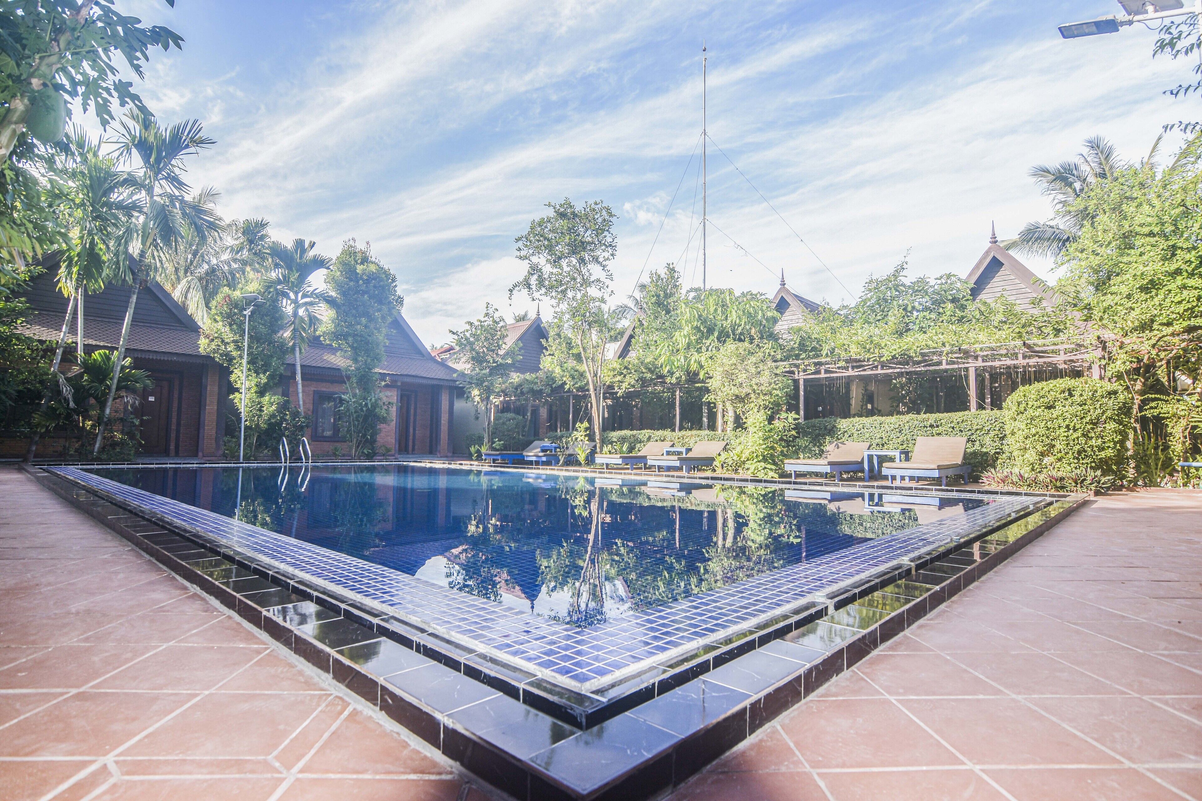 Une piscine rectangulaire bordée de carreaux, entourée de verdure et de bâtiments de style traditionnel.