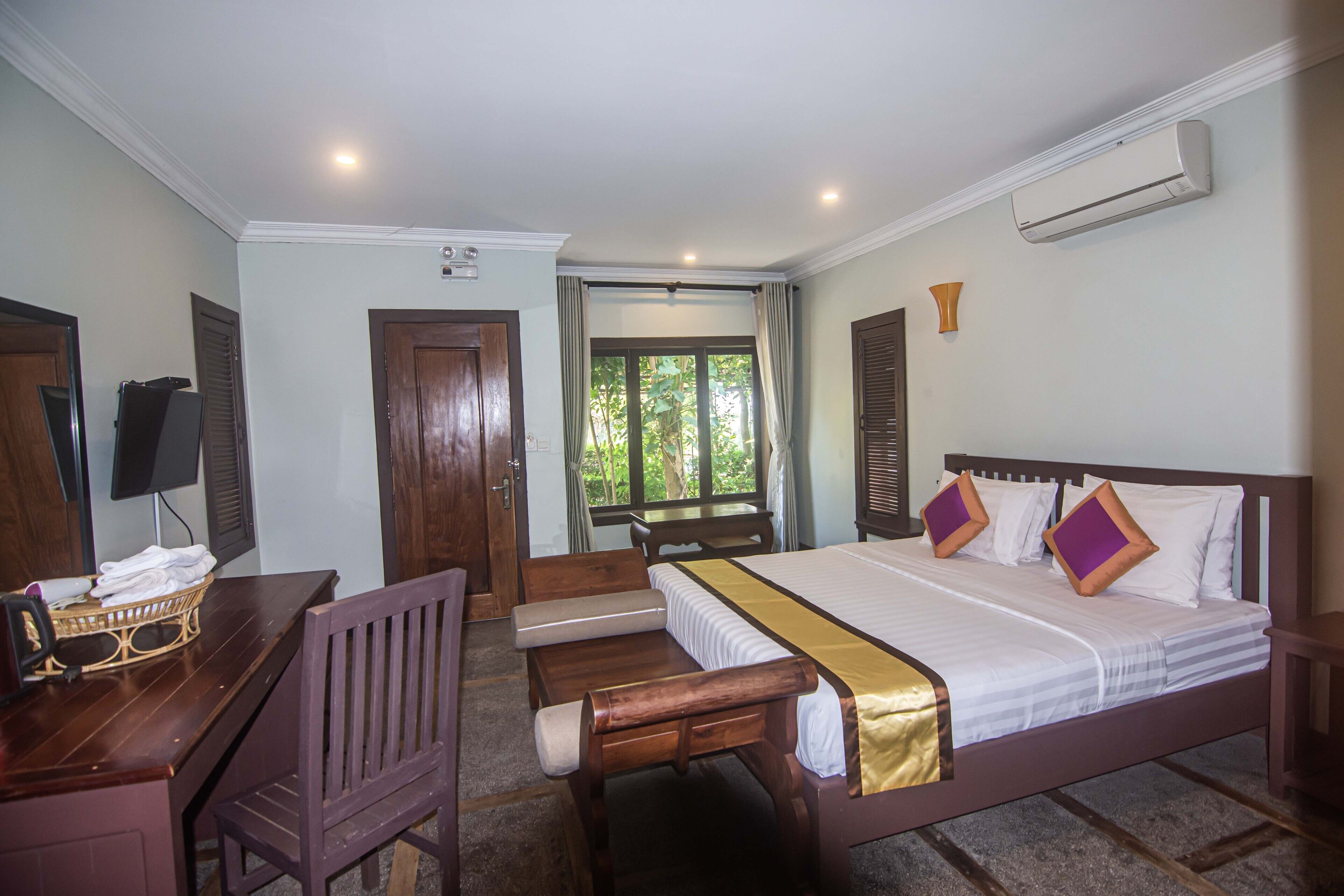 A hotel room with a bed, a desk, a chair, a window with curtains, and an air conditioning unit.