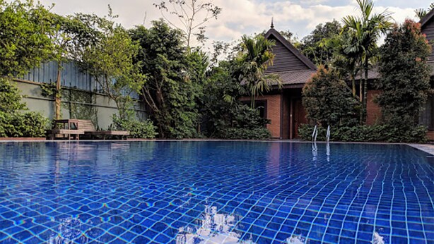 Una piscina con cubierta de baldosas, rodeada de exuberante vegetación y un edificio tradicional con techo de tejas.