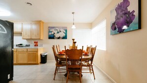 A dining area with a wooden table and chairs, a kitchen with wooden cabinets, and two wall-mounted flower paintings. - glorious sunsets  (Seaside)