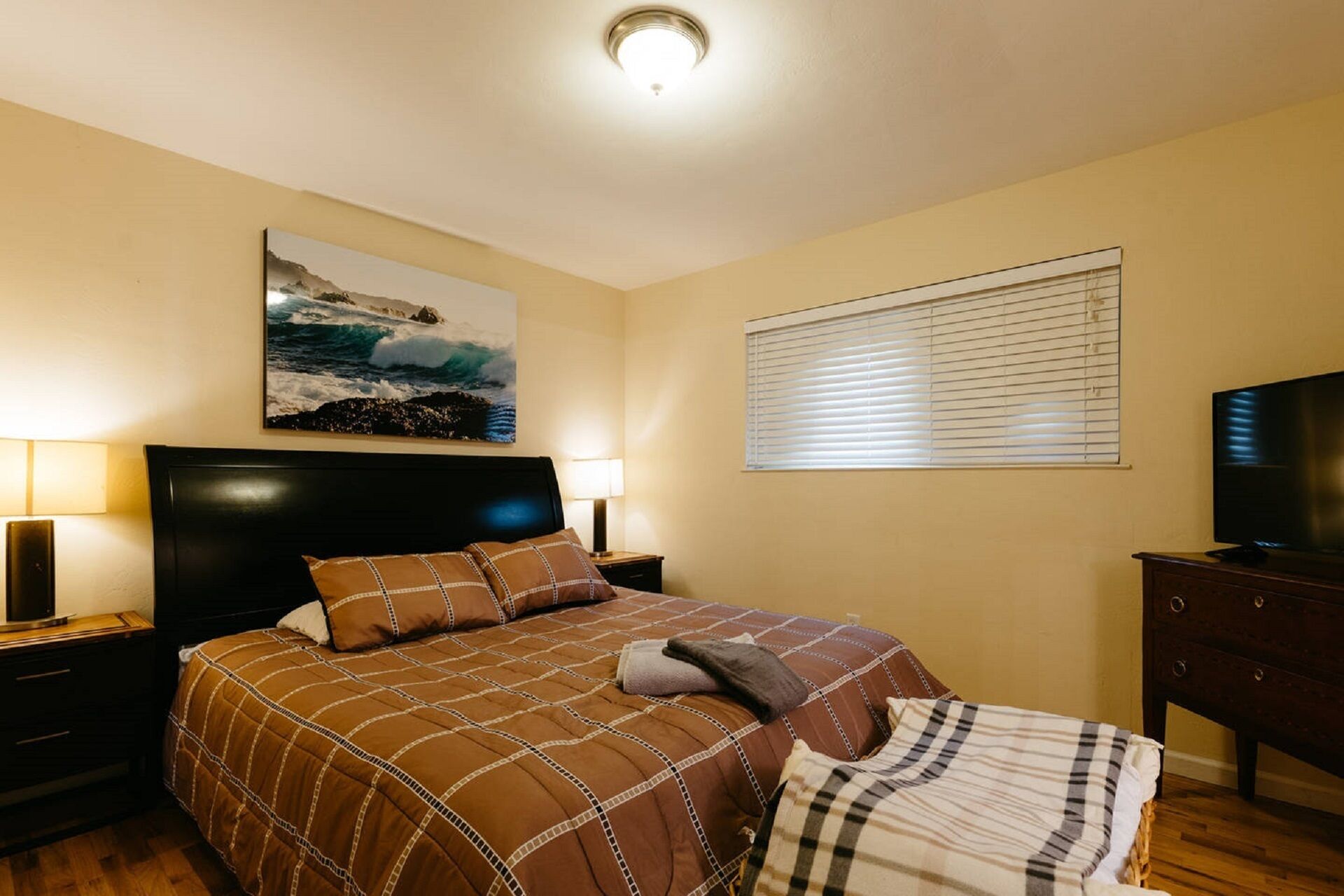 A bedroom with a bed, two bedside tables, a television, a window with blinds, and a painting on the wall.
