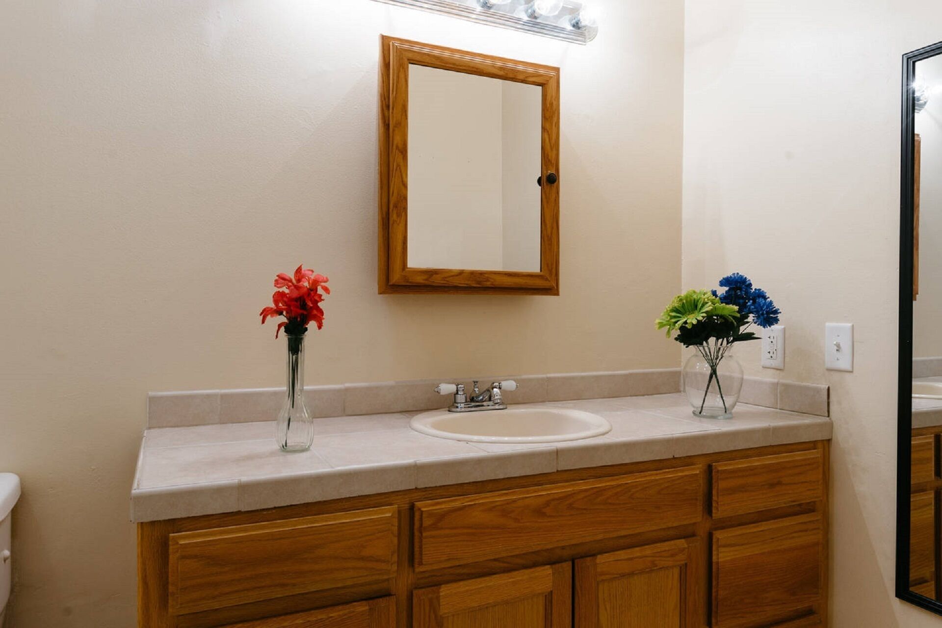 Un baño con tocador de madera, un lavabo blanco y dos jarrones con flores.
