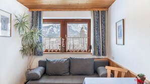 Een kamer met een houten plafond, een bank met kussens, een tafel met een witte tafelkleed, een potplant en een raam met gordijnen.