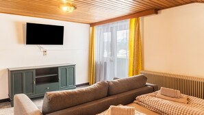 Una habitación con techo de madera, televisor montado en la pared, un sofá, una cama con colcha a cuadros y una ventana con cortinas amarillas.
