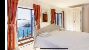Een slaapkamer met een groot bed, een balkon met gordijnen en uitzicht op zee, plus een houten deur.