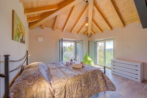 Un dormitorio con una cama grande, vigas de madera en el techo, un ventanal con vistas y un aparador blanco.