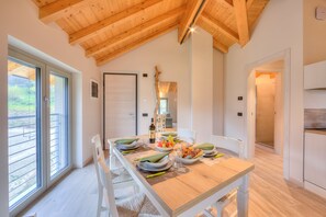 A dining area with a wooden table set for four, a view of the outdoors through large sliding doors, and a kitchen area visible through an open doorway. - Ricomilla-Finestra Sul Verde- priv park, Roè Volciano, Italy (Roè Volciano)