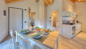 Ein Essbereich mit Holztisch, gedeckt für vier Personen, eine Küche mit modernen Geräten und eine hohe Decke mit freiliegenden Balken.