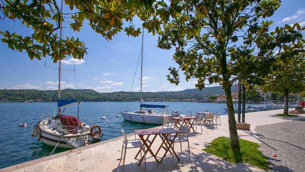 Ein Uferbereich mit vertäuten Booten, Außenplätzen und Blick auf Wasser und Hügel im Hintergrund.