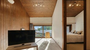 A modern room with a flat-screen TV on a wooden stand, a wicker chair, and a bed with a view of mountains through large windows. - La Maison De Montroc Family-Holidays w/4BR, + Fireplace, Sleeps 8, Le Tour (Argentière), France (Le Tour (Argentière))