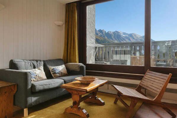 Un salon avec un canapé, une table en bois et une chaise, une grande fenêtre donnant sur les montagnes et une balustrade de balcon.