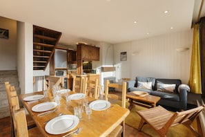 Amplia sala de estar con una mesa de comedor de madera dispuesta para cuatro personas, un sofá y una escalera que conduce a una cocina.