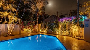 A swimming pool with a tiled deck, surrounded by palm trees and a thatched-roof building.