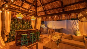 An outdoor seating area with a thatched roof, wooden furniture, and a bar with stools.