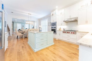 A modern kitchen with white cabinetry, a central island, and a dining area with a wooden table and chairs. - Honey's Haven | Luxury St. Simons Island Vacation Rental with Private Pool Near Pier Village (St. Simons Island)