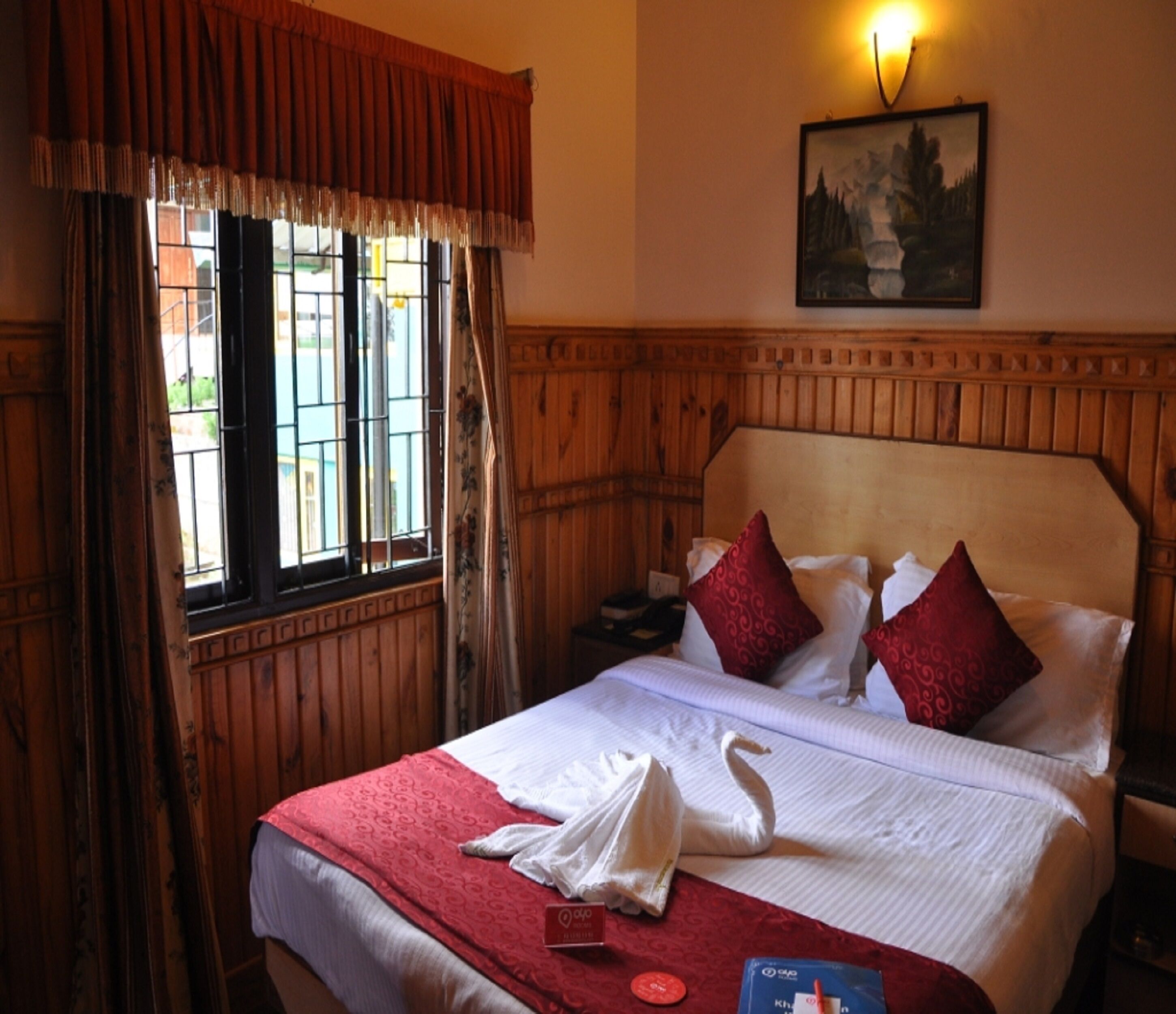 Ein Hotelzimmer mit einem hölzernen Kopfteil, einem Bett mit weißer und roter Bettwäsche, einem Fenster mit Vorhängen und einem Bild an der Wand.