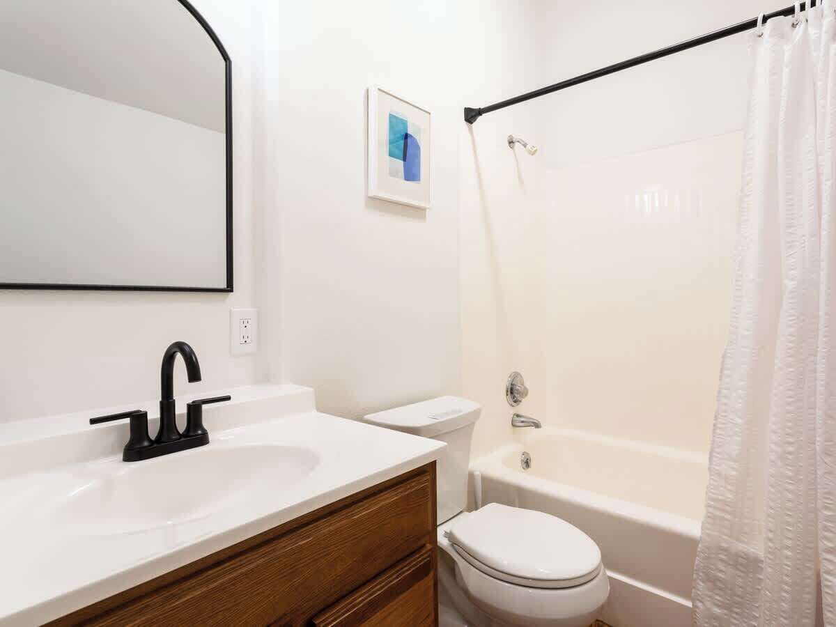 Une salle de bain avec un lavabo blanc, un robinet noir, des toilettes blanches et une baignoire agrémentée d’un rideau de douche blanc.