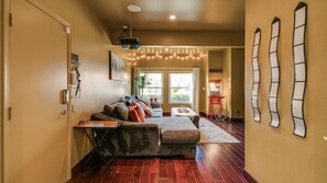 A living room with a sofa, a coffee table, and a TV. There are string lights on the ceiling and a door with a sign.