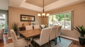 A dining room with a wooden table, beige chairs, a chandelier, a large mirror, and a view of the outdoors through a window.