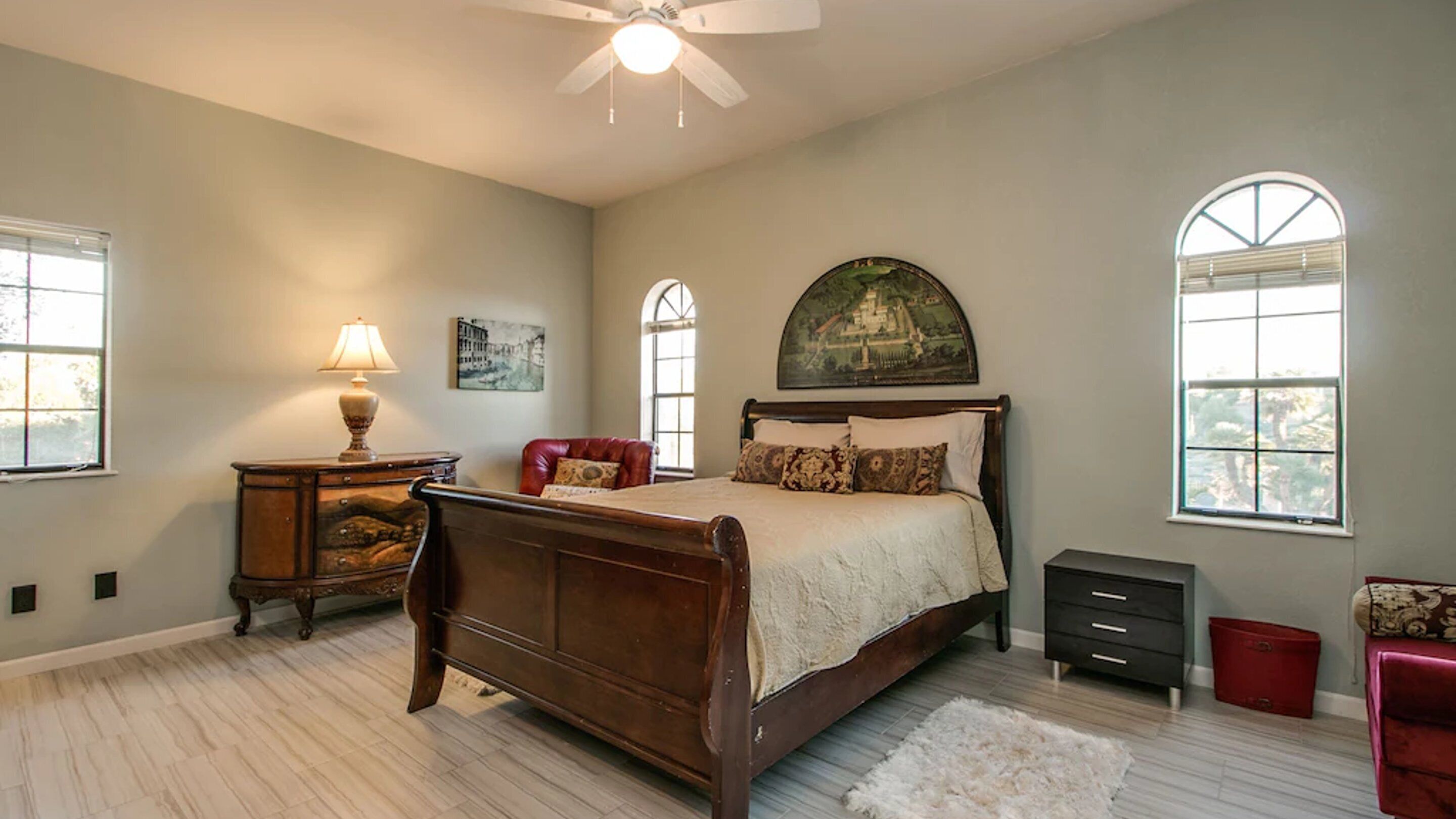 Une chambre à coucher avec un lit en bois, une table de chevet, une lampe, un fauteuil rouge, une commode et une grande fenêtre donnant sur un beau paysage.